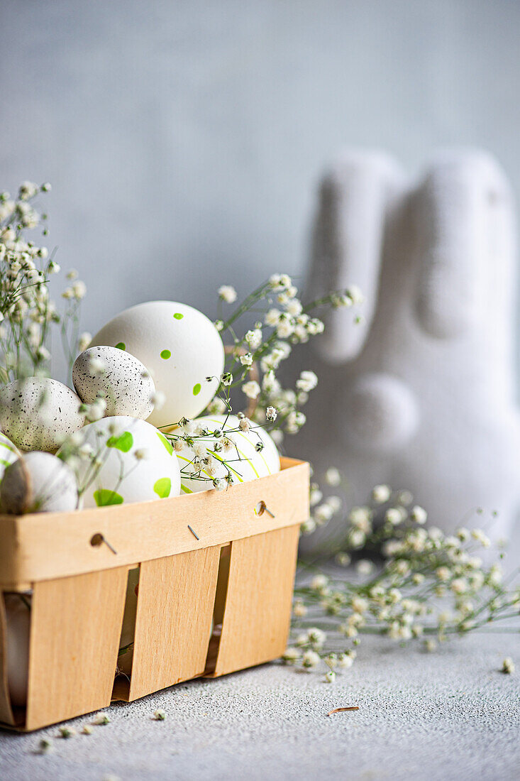 A selection of decorated Easter eggs nestled in a wooden basket alongside sprigs of gypsophila, with a soft gray background