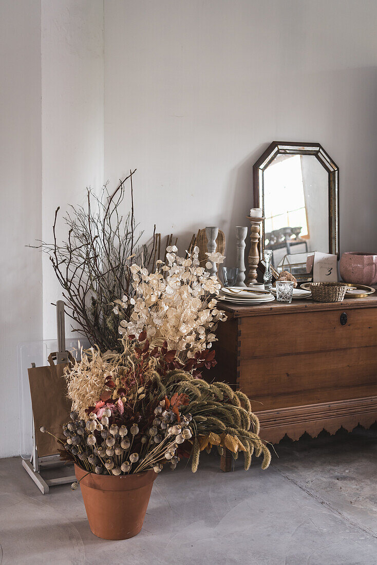 Innenansicht eines hellen Raums mit einem Holzschrank und einem Spiegel in der Nähe von trockenen dekorativen Blumen in einem Topf an einer weißen Wand