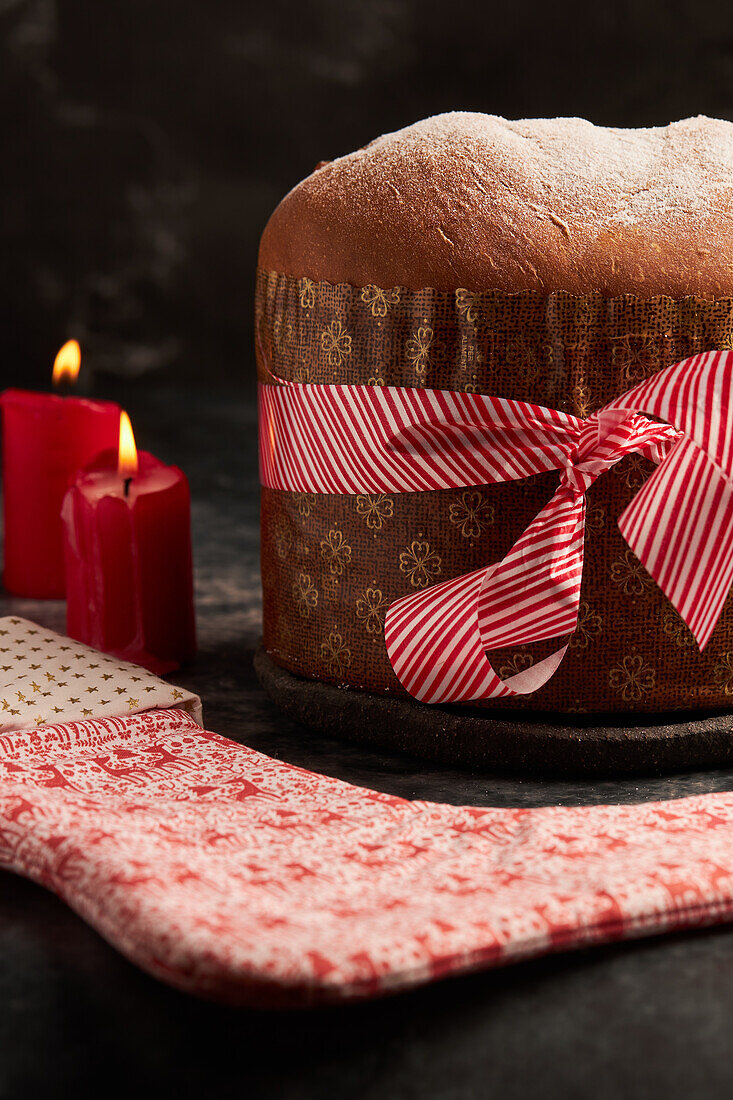 Ein wunderschön präsentiertes Panettone-Brot, eingewickelt mit einem gestreiften roten Band, begleitet von einem dekorativen Stoff mit festlichen Mustern, inmitten von Kerzenlicht