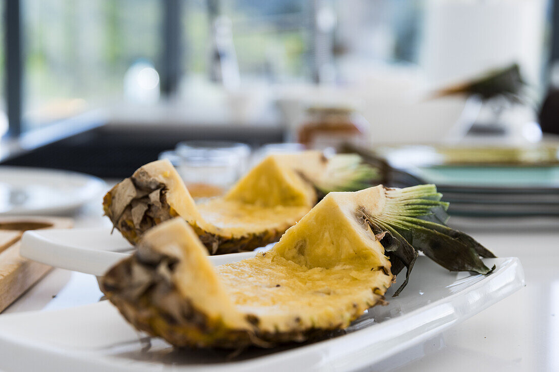 Appetitlich geschnittene frische Ananas mit dorniger Schale und grünen Blättern auf einem weißen Teller auf dem Küchentisch, Eierstock und Kern entfernt, vor unscharfem Hintergrund