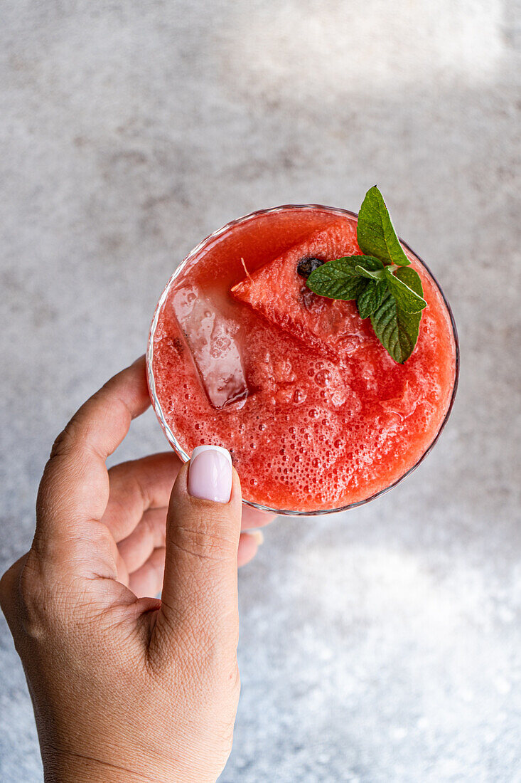 Top view of crop person touching glass of fresh Margarita cocktail with watermelon smoothie covered with mint leaves