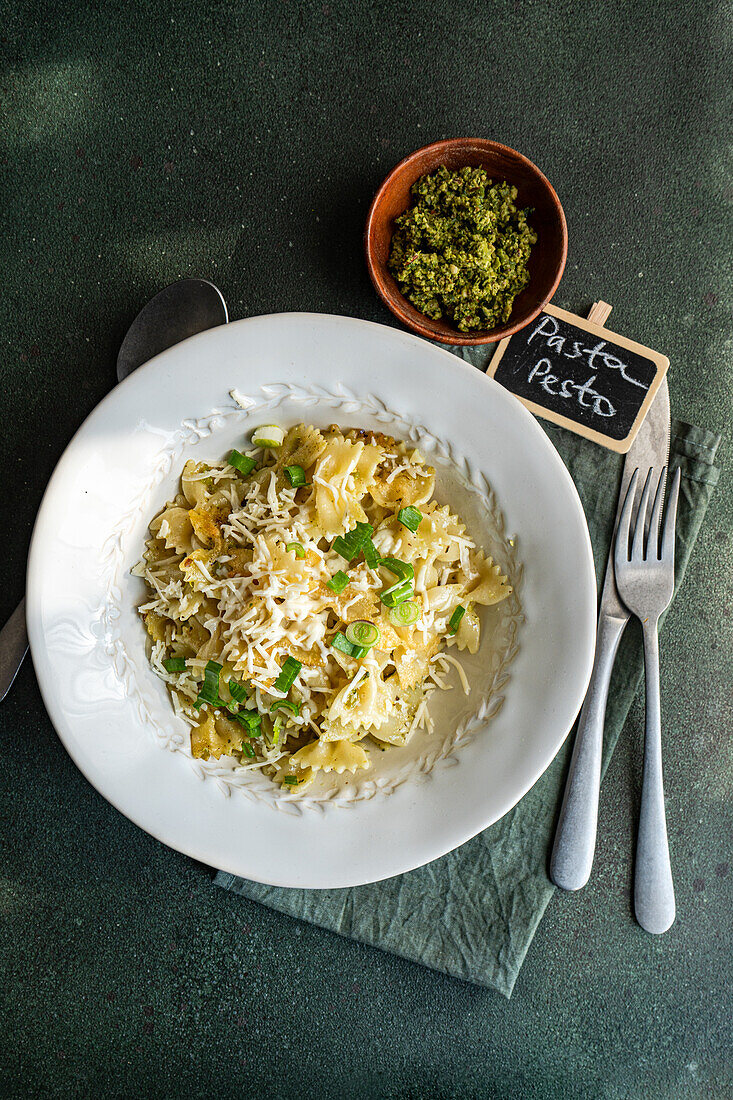 Overhead shot of a delicious bowl of pesto Farfalle pasta with grated cheese, on a dark background with a small bowl of chopped herbs and a label indicating the recipe