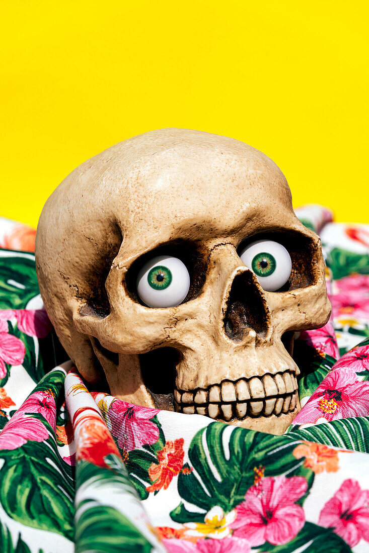 Spooky human skull looking at camera with green eyes while being placed on fabrics with drawn flowers and yellow background