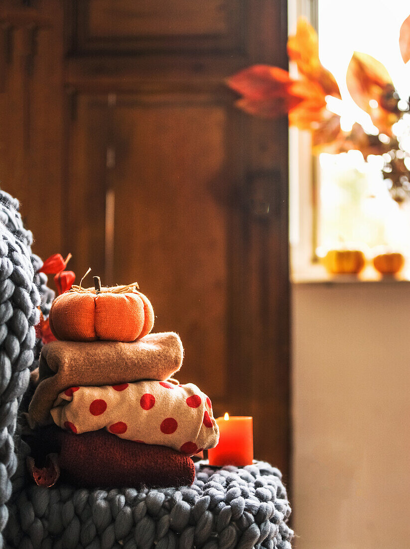 Autumn still life with folded cozy sweaters, wool blanket, pumpkin, candle at window background with sunlight. Warm clothes and seasonal interior decoration at home. Front view