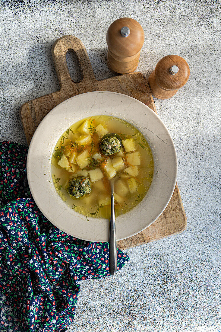 From above bowl of savory meatball soup with diced vegetables, presented on a rustic wooden cutting board, paired with a patterned napkin and seasoning mills.