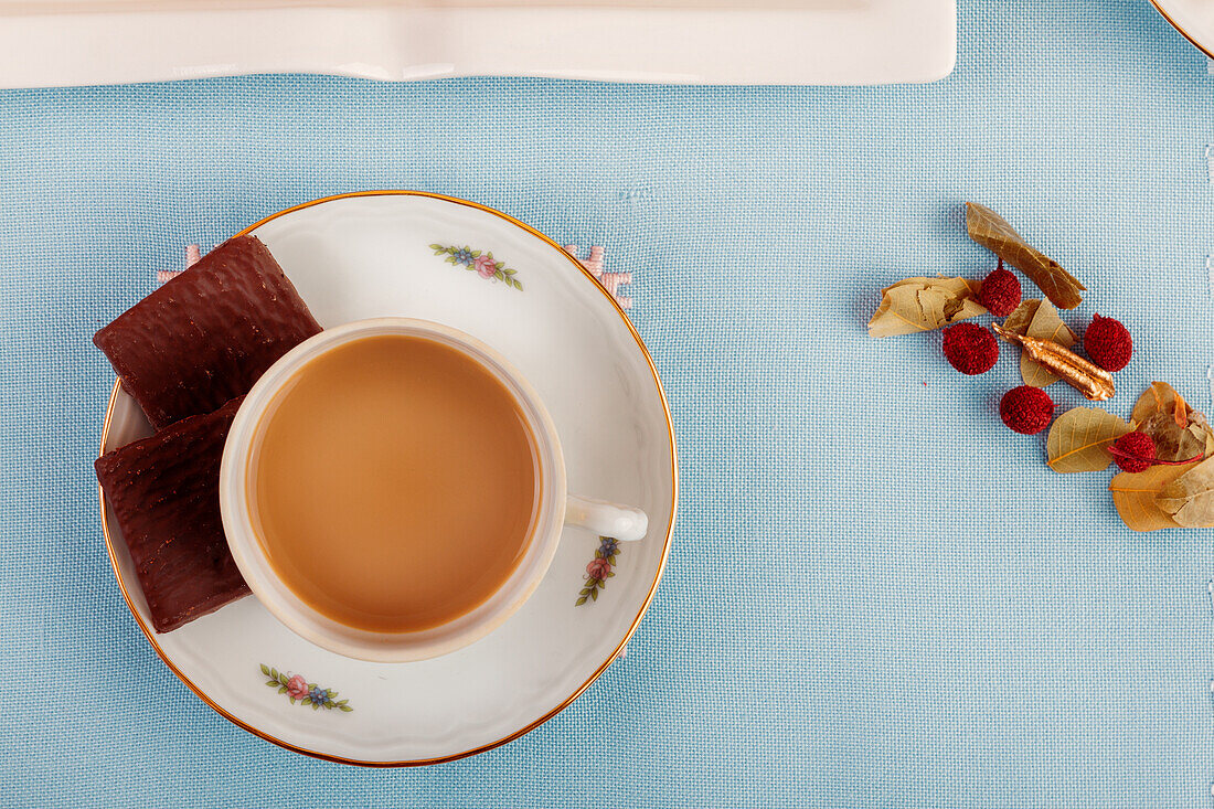 Eine Tasse Tee mit Milch, serviert auf einer Untertasse neben Schokolade und Nüssen auf blauem Hintergrund