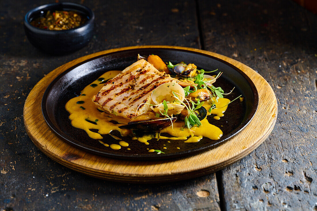 High angle of traditional Ecuadorian dish appetizing grilled fish steak garnished with mussels herbs and yellow sauce served on black plate