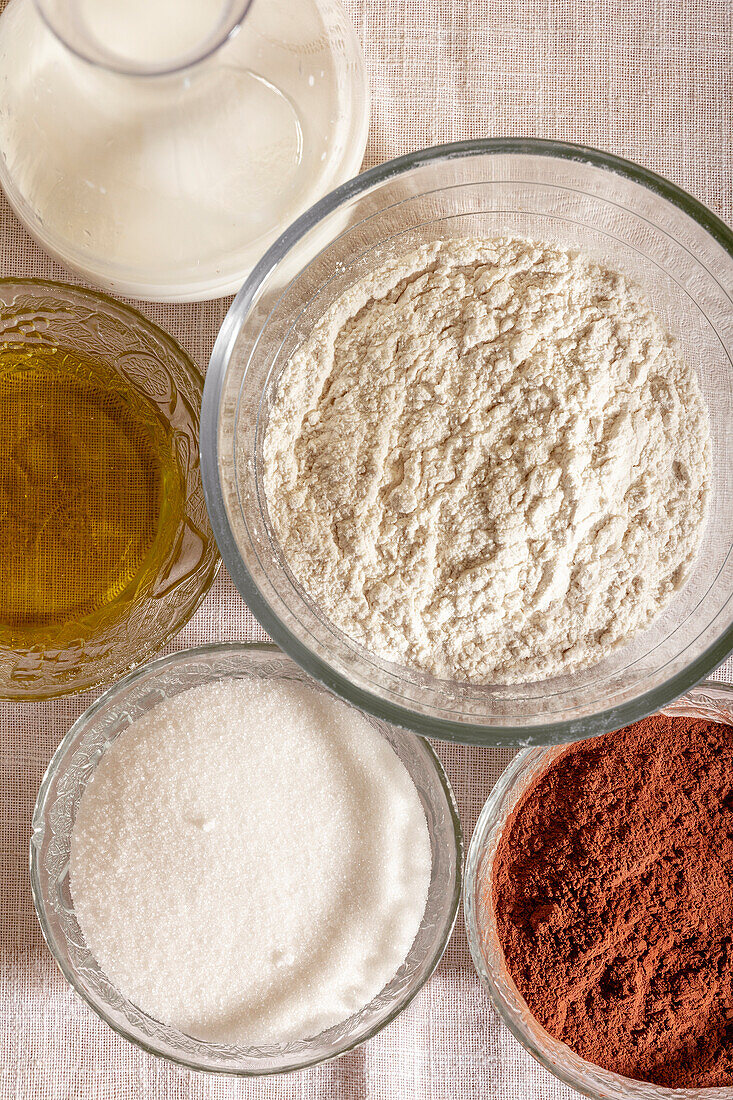 Overhead view of glass bowls filled with flour, sugar, cocoa, oil, and milk, ready for baking placed on table