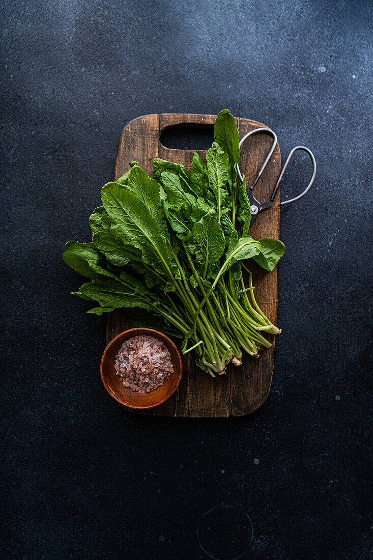 Top view of bunch of fresh arugula leaves alongside a bowl of pink salt and scissors on a rustic wooden cutting board, set against a dark concrete tabletop