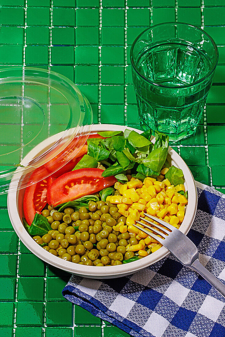 Hochformatige Salatschüssel mit Tomatenscheiben, Spinatblättern, Maiskörnern und Erbsen auf einer Serviette auf einer grünen Fläche mit einer Gabel neben einem Glas Wasser
