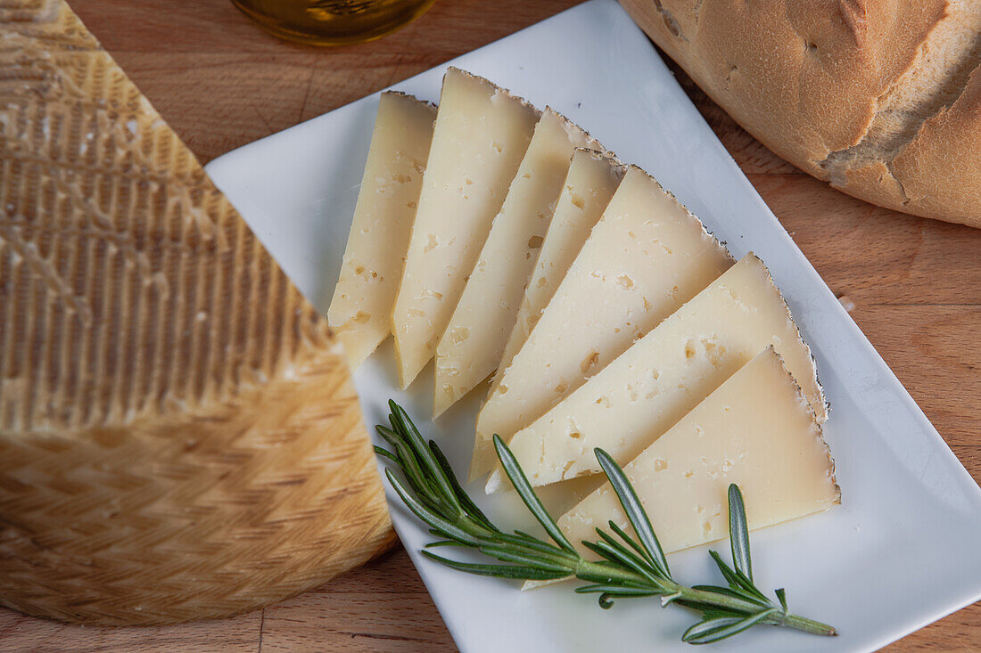 Sliced artisan cheese on a white plate accompanied by fresh rosemary sprigs and a loaf of bread, with olive oil in the background.