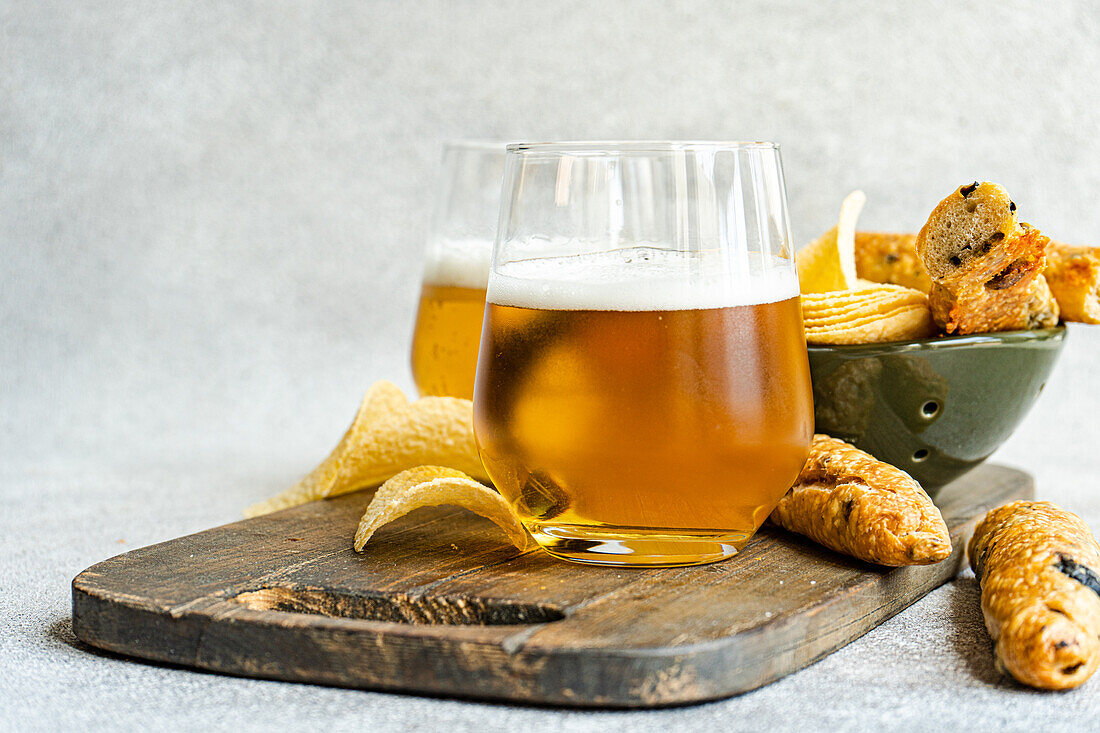 Tasty pieces of crisp potato chips in bowl with bread sticks and glasses of beer on wooden board placed over table in daylight