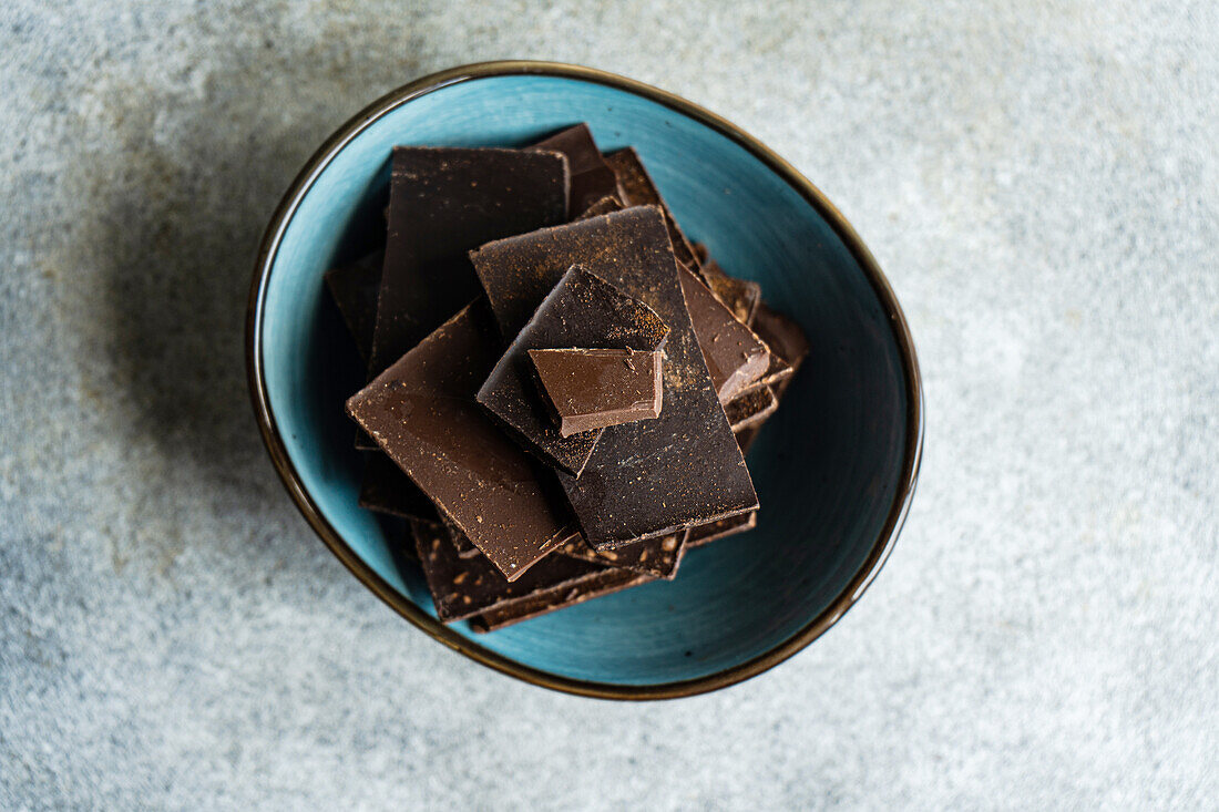 Top view of pile of pieces of different kinds of chocolate placed in crop ceramic bowl on gray blurred table