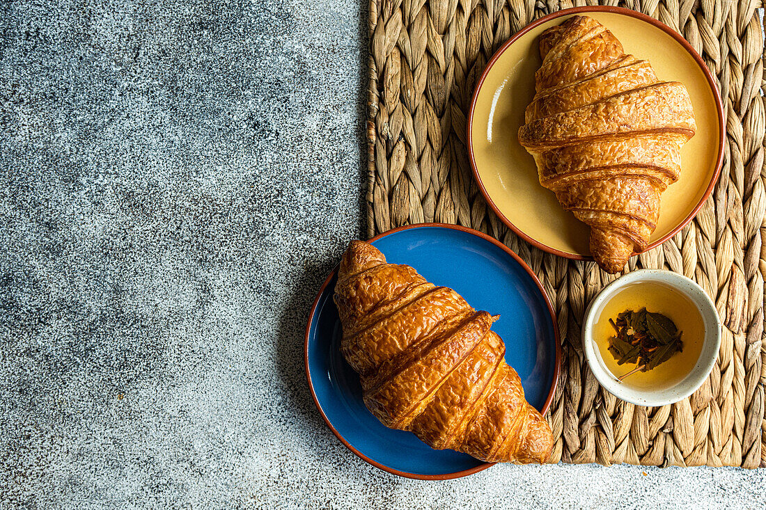 Draufsicht auf frisch gebackene Croissants auf bunten Keramiktellern auf brauner Serviette neben einer Tasse grünem Tee vor grauem Hintergrund