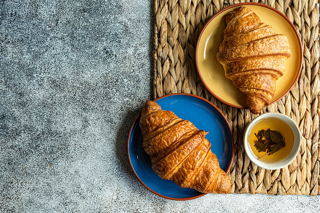 Top view of fresh baked croissants on the colorful ceramic plates placed on brown napkin near cup of green tea against gray background
