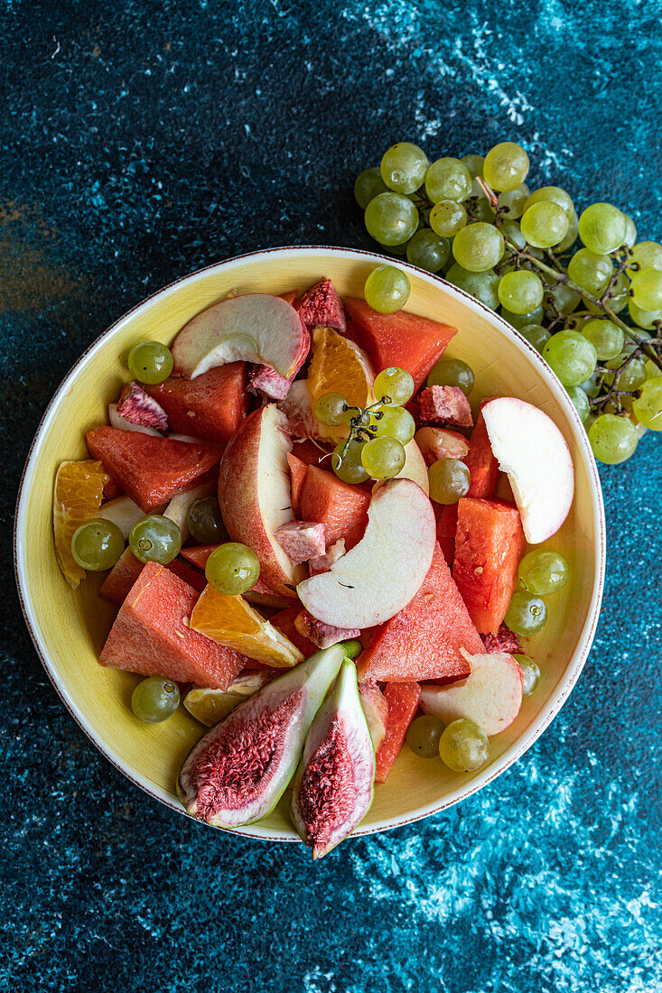 Top view of ceramic bowl of slices of different fruits placed on blue surface near blurred grapes