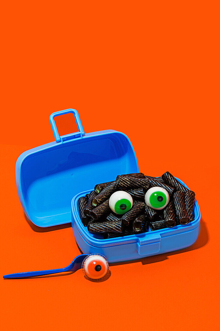 Horror-Lunch mit schwarzen Nudeln und Augen in Lunchbox auf orangem Hintergrund neben Gabel mit Auge
