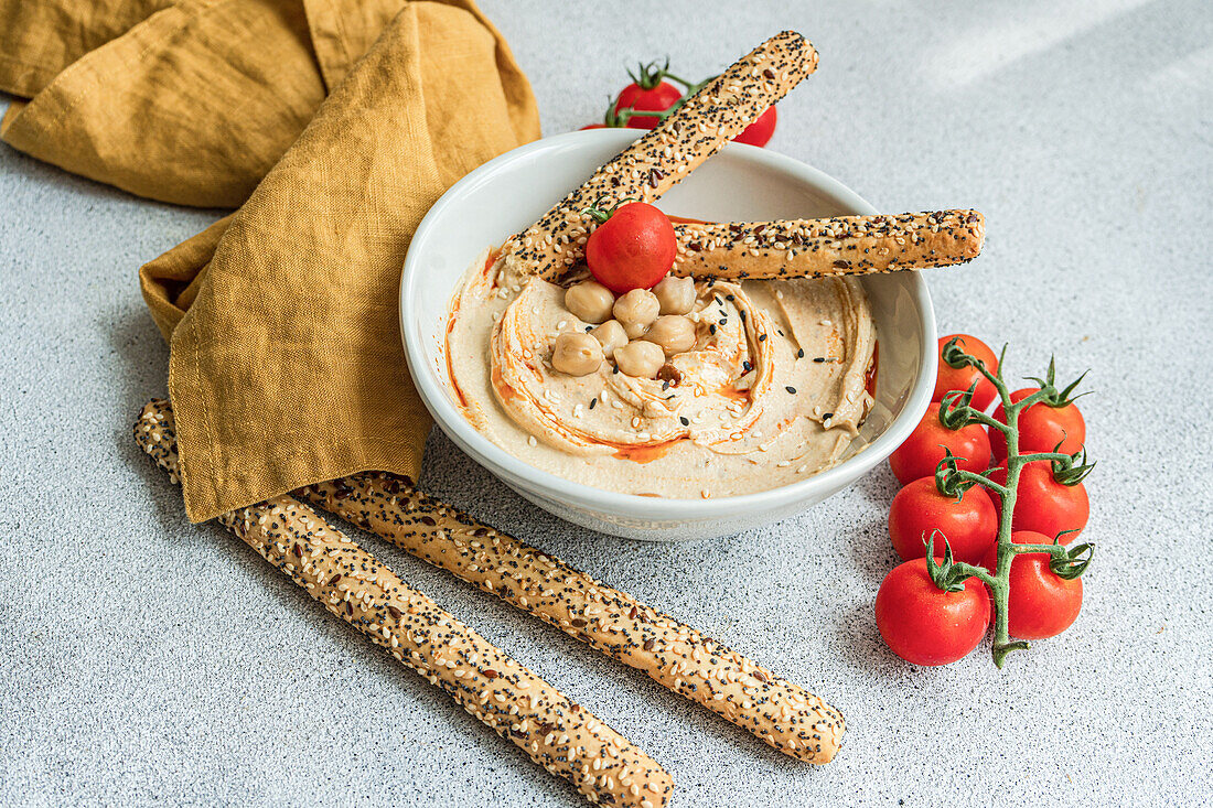 Hochformatiger gesunder Pflanzenteller mit Hummus und Tomaten, serviert mit Brotstäbchen in einer Schale neben einer Serviette vor unscharfem Hintergrund