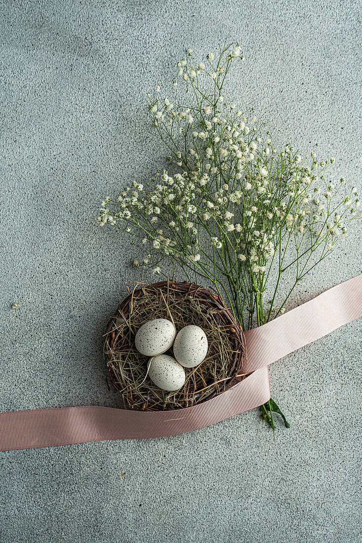 Draufsicht auf gesprenkelte Ostereier in einem rustikalen Korb, verziert mit zartrosa Schleife und zarten weißen Blumen, arrangiert auf einem gesprenkelten grauen Hintergrund