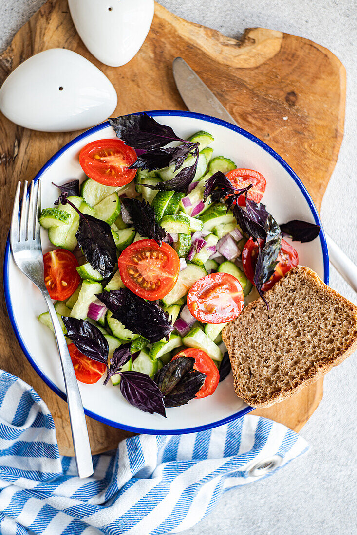 Top view of Seasonal vegetable salad with organic cucumber, tomato, onion and red basil leaves served on plate placed on wooden tray near napkin against gray background