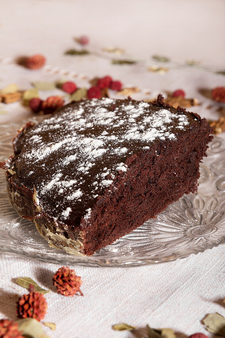 High angle of tempting slice of rich chocolate cake sprinkled with powdered sugar, presented elegantly on a glass plate with a decorated table background