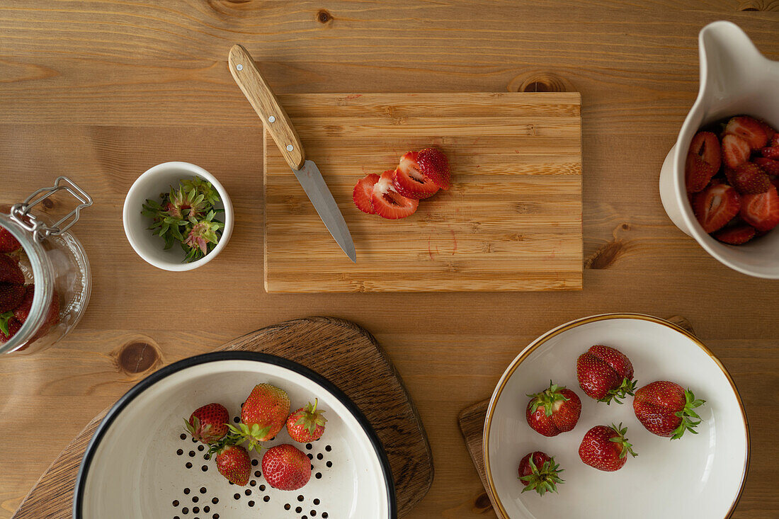 Draufsicht auf frisch geschnittene Erdbeeren auf einem Holzbrett und in einer Schüssel mit Messer auf einem Holztisch bei der Zubereitung von Erdbeermarmelade in einem Haus