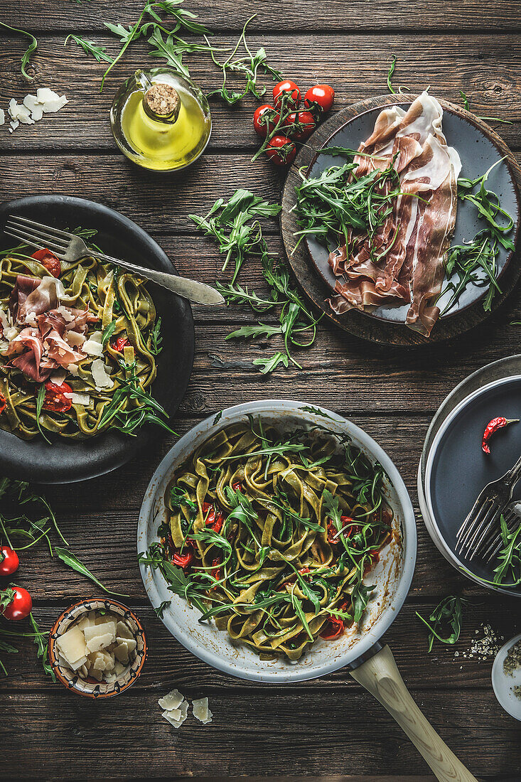 Italian food background with green cooked pasta in cooking pan with parma ham, arugula, tomatoes and cheese on rustic wooden kitchen table. Homemade delicious food on plates. Ready to eat. Top view.