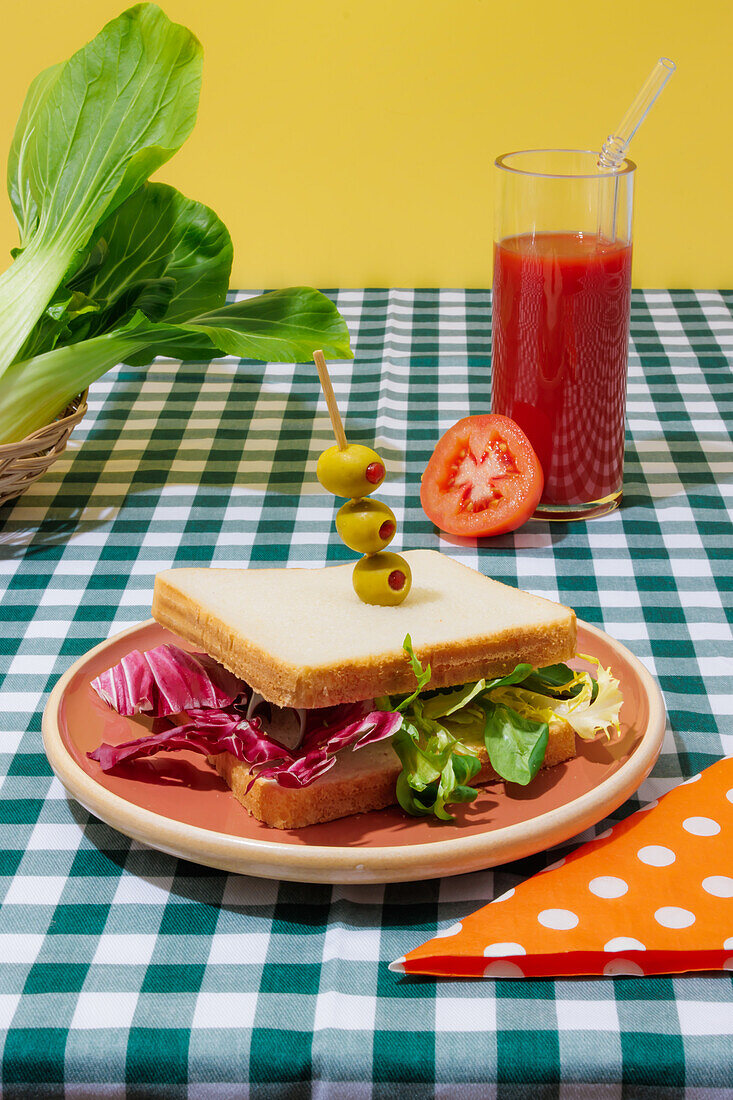 Appetizing healthy sandwich with fresh salad served on plate with olives near glass of tomato juice with glass straw on checkered tablecloth