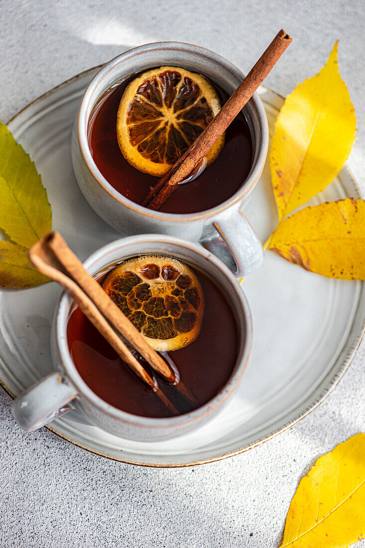 Von oben auf zwei Tassen mit aromatischem Gewürztee mit Zimtstangen, Anis und getrockneten Orangenscheiben, umgeben von leuchtend gelben Herbstblättern auf einem strukturierten grauen Hintergrund