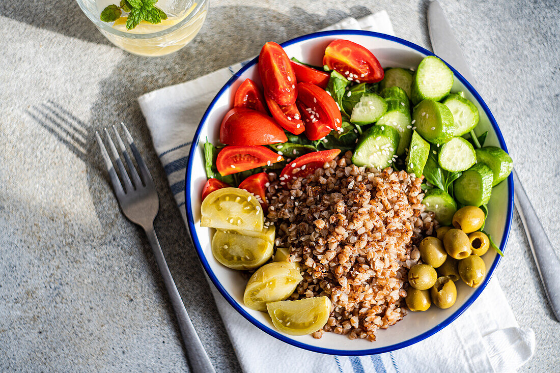 Top view of healthy lunch bowl with boiled organic buckwheat, fresh cucumber and tomato, and fermented tomato and olives served with glass of pure water with lemon, ice and mint against blurred gray surface