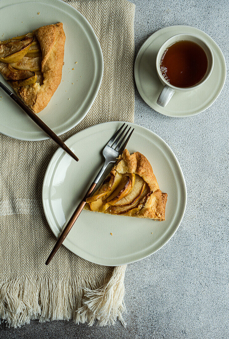 Draufsicht auf Teller mit Stücken von Apfelkuchen und Gabeln auf Serviette neben Tasse Tee vor grauem Tisch