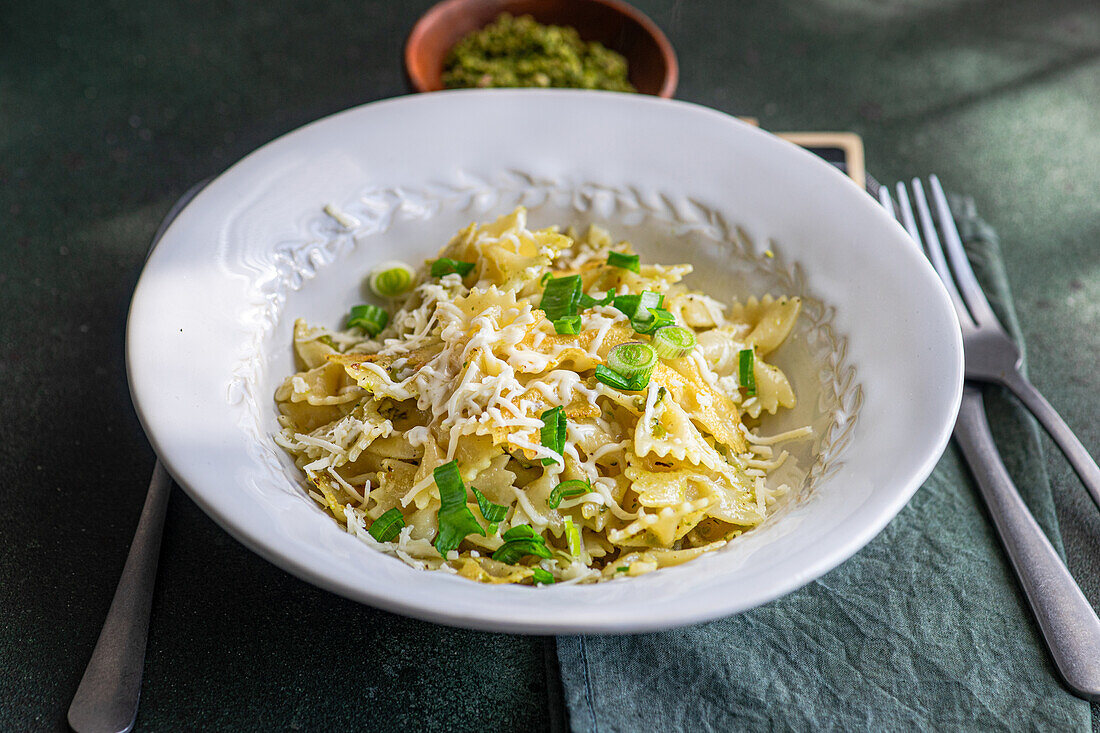 A white bowl filled with Farfalle pasta, peas, and grated cheese, accompanied by a pesto sauce container, placed on a dark surface with cutlery alongside.