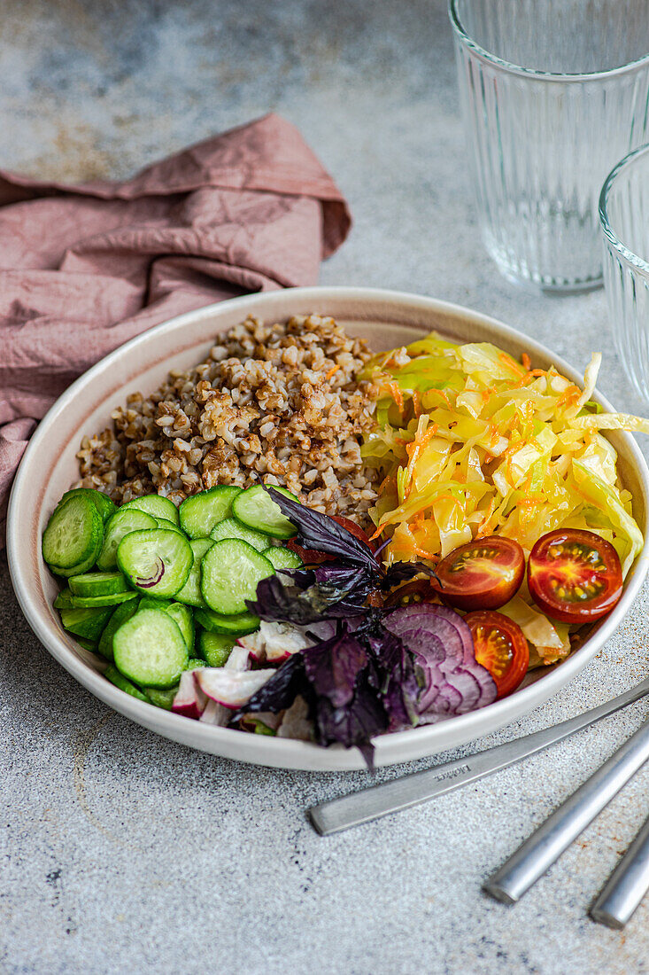 High angle of bowl with delicious buckwheat containing sliced fresh basil plant with cabbage and cucumber with cutlery and glasses placed on gray table with napkin