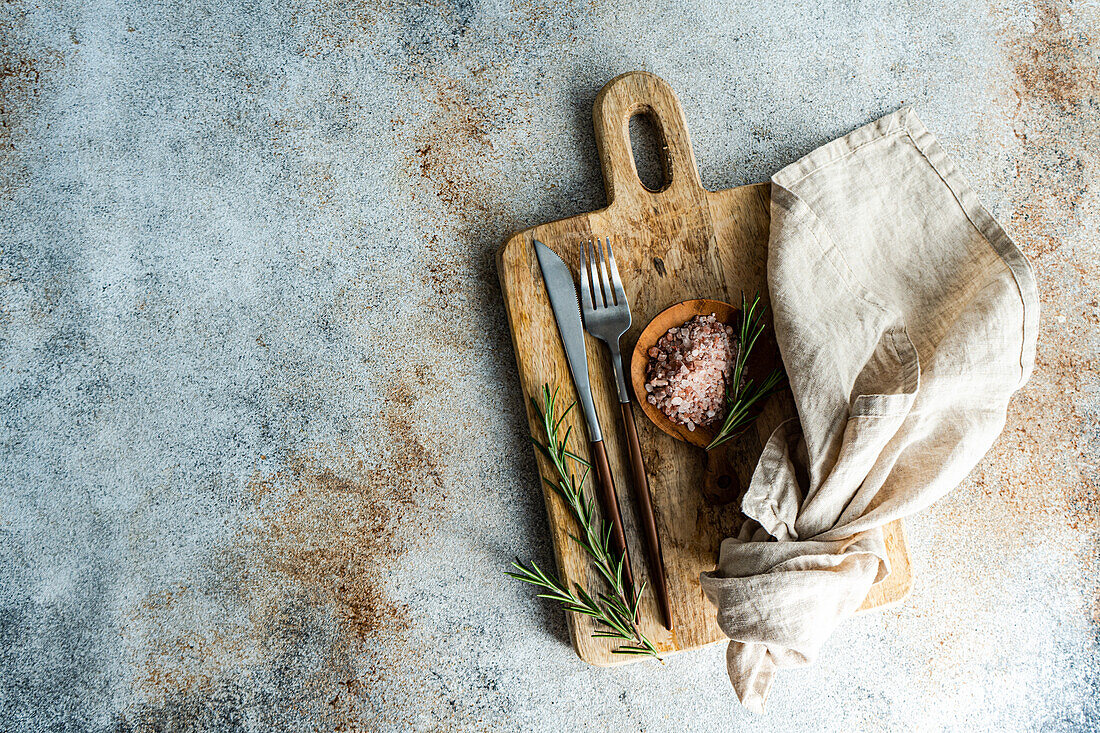 From above rustic kitchen setup on wooden cutting board with a fork, knife, a sprinkle of pink Himalayan salt, fresh rosemary sprigs, and a linen napkin, laid out on a textured concrete background