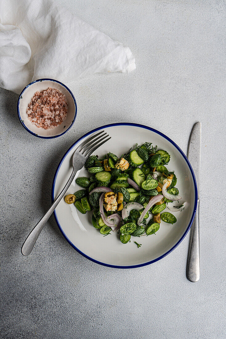 Top view of cucamelon salad with red onion, olives and spicy cheese served on white plate with fork and knife near napkin on gray table in daylight