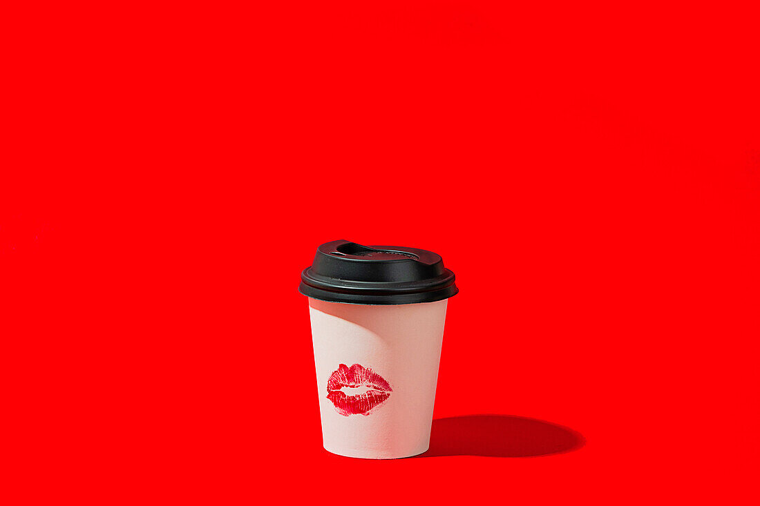 Ein Papierkaffeebecher mit schwarzem Deckel und einem Lippenstiftfleck steht vor einem leuchtend roten Hintergrund, der einen starken Kontrast hervorhebt