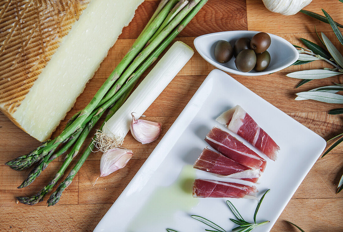 Gourmet selection of Manchego cheese, green asparagus, sliced prosciutto, olives, and garlic on a wooden table.