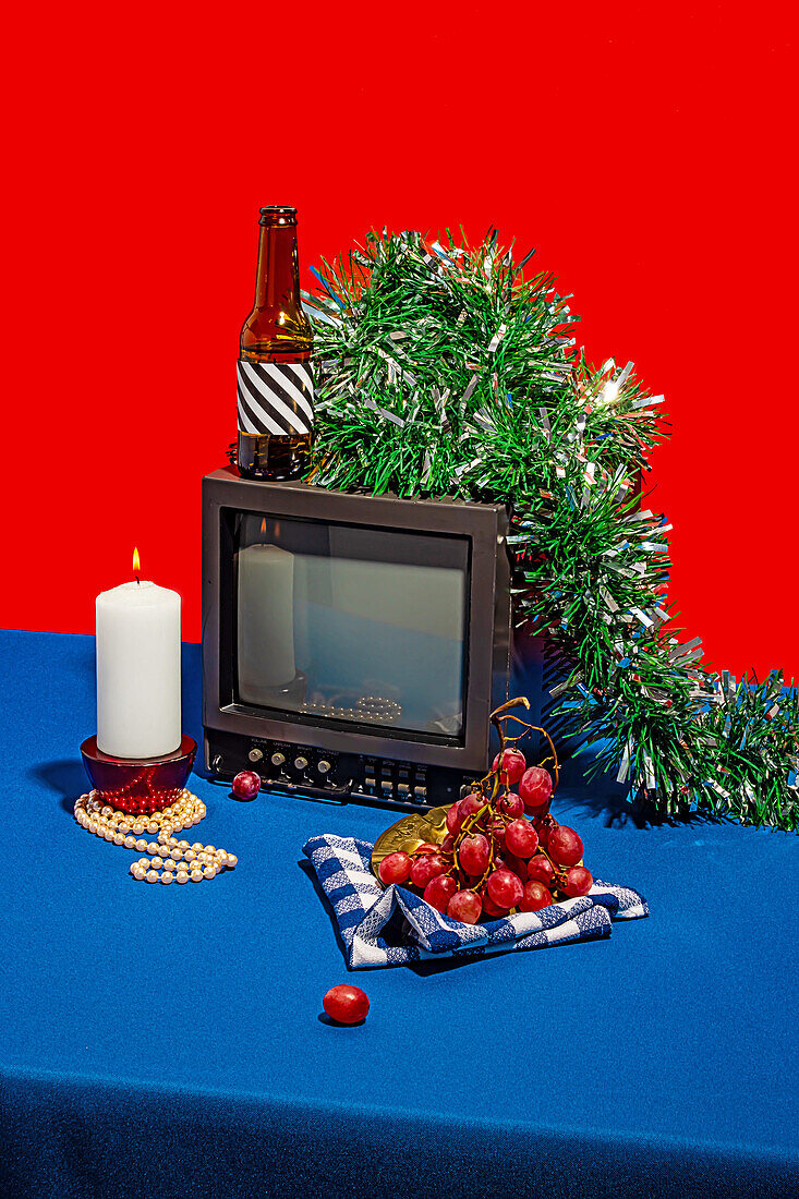 High angle of vintage television set surrounded by an array of objects, including a bottle with a striped label, fresh grapes on a checkered cloth, a white candle and green tinsel, all set against a red backdrop on a blue table