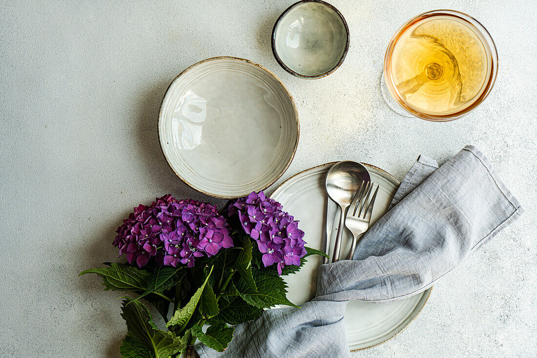 Draufsicht auf eine lila Hortensie, die auf einem weißen Tisch neben Keramiktellern und einem Glas mit Getränk steht