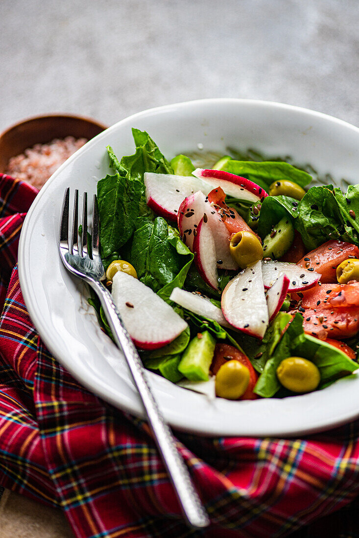 Close-up of healthy vegetable salad with fresh organic spinach leaves, juicy tomatoes, crunchy radish slices, and tangy olives served on a white plate with fork on the side all placed on textured surface with checkered cloth