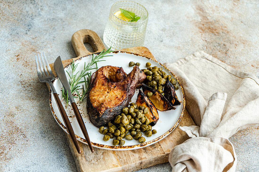 Close up of healthy grilled salmon steak seasoned with spices next to roasted lemons and capers on a wooden board with a linen napkin and lemon water