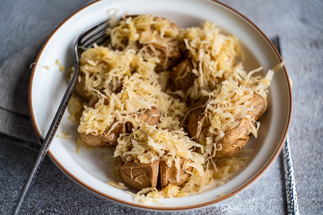 Draufsicht auf einen Teller mit Pellkartoffeln und fermentiertem Krautsalat, serviert mit Gabel und Messer, auf einer grauen, strukturierten Oberfläche mit einer gefalteten Stoffserviette