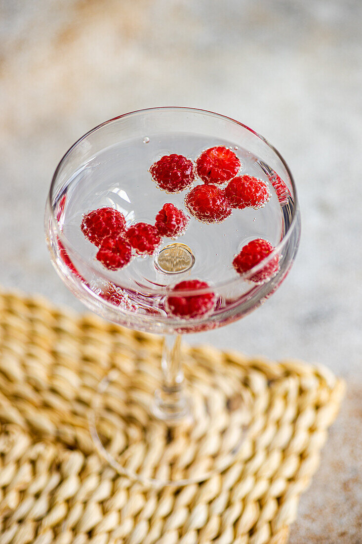 Hoher Winkel eines transparenten, glatten Glasbechers, gefüllt mit erfrischendem Cocktailsekt mit reifen, frischen Himbeeren, der auf einer Tischdecke auf einem Marmortisch steht, vor einem verschwommenen Hintergrund