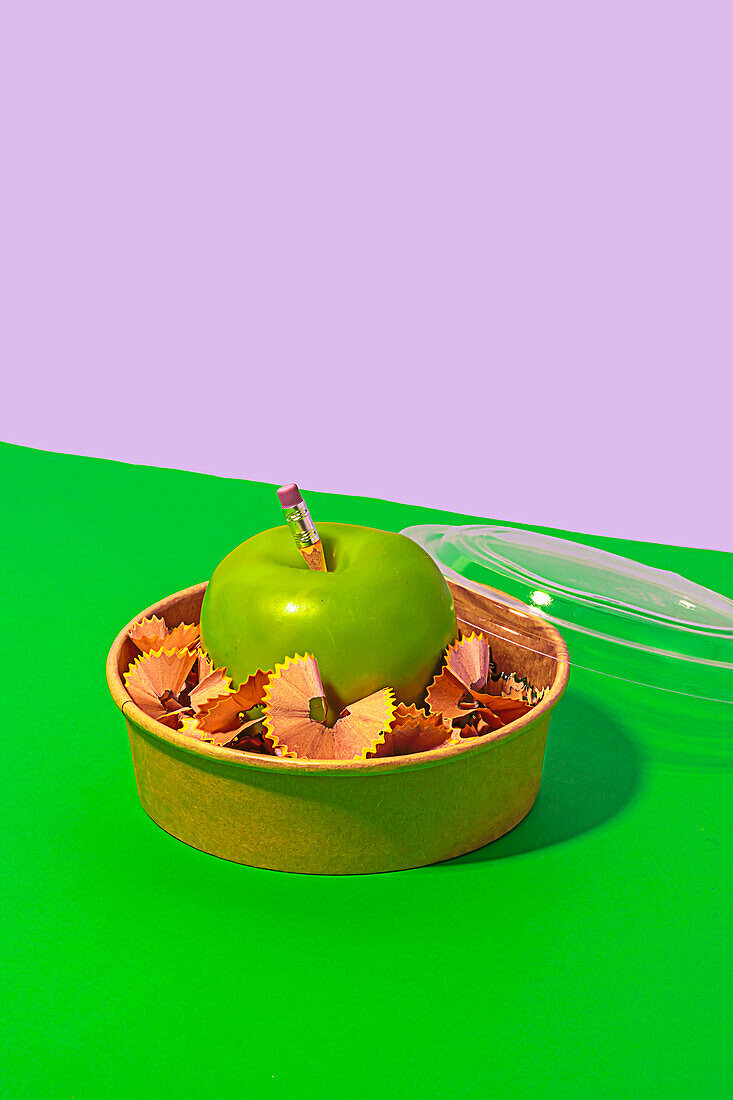 From above of healthy apple surrounded with pencil shavings in lunch box placed on green and white background representing concept of zero waste