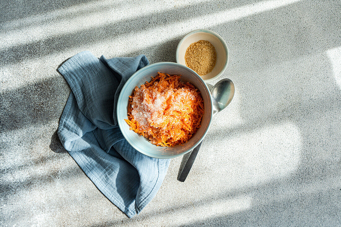 Top view of a light blue bowl containing freshly grated carrot sprinkled with sugar, placed next to a gray cloth, a small beige bowl with brown sugar, and a metallic spoon, all set against a textured gray background with sunlight casting soft shadows