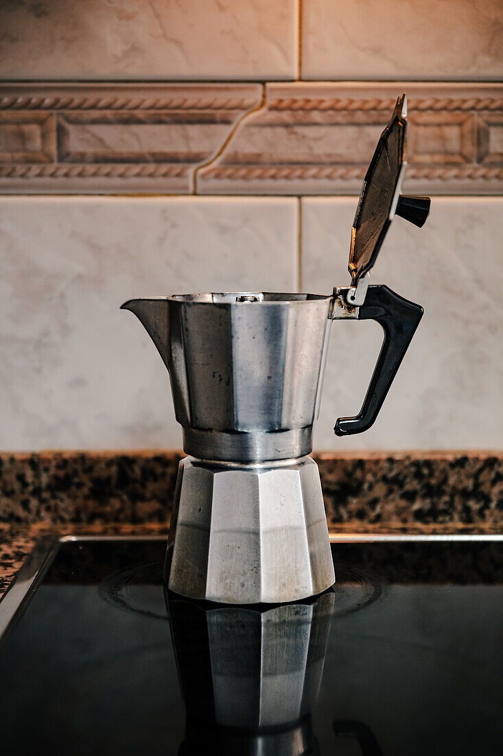 Close-up of stainless steel stovetop espresso maker placed on sleek cooktop with an ornate tiled backsplash providing a contrasting backdrop emphasizing the allure of home brewed Italian coffee
