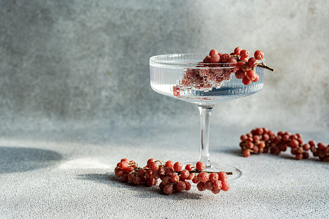 An elegant cocktail glass filled with wine perched atop a textured grey background, surrounded by scattered bunches of red grapes