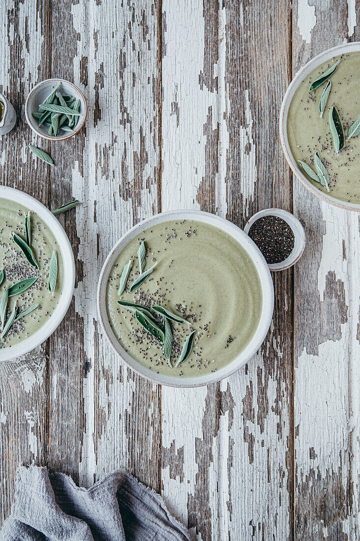 Ceramic bowls of green cream soup with sage served with seeds and herbs on wooden table with shabby surface