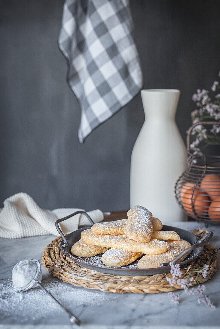 Tray with crunchy ladyfingers finger sponge cake placed on table near eggs and jar on gray background in kitchen at home