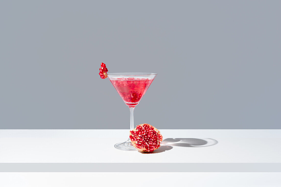 Glas gefüllt mit rotem Granatapfel-Cocktail mit Granatapfelkernen neben einem saftigen frischen Granatapfel, vor einem grauen Hintergrund, der einen weichen Schatten wirft
